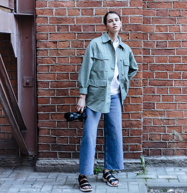 picture of a woman standing in front a wall on a sidewalk holding a camera and wearing stylish clothes with comfortable sandals  